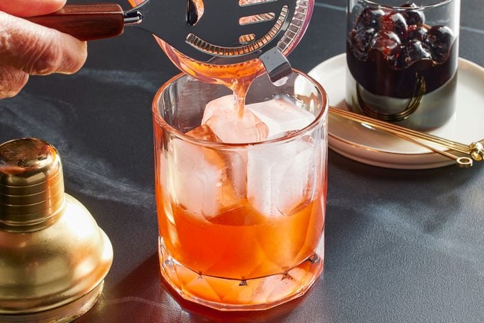 Eye Level horizontal image of an orange liquid being strained into a glass filled with ice. A shaker is visible to the left and a glass of cherries is visible in the background.