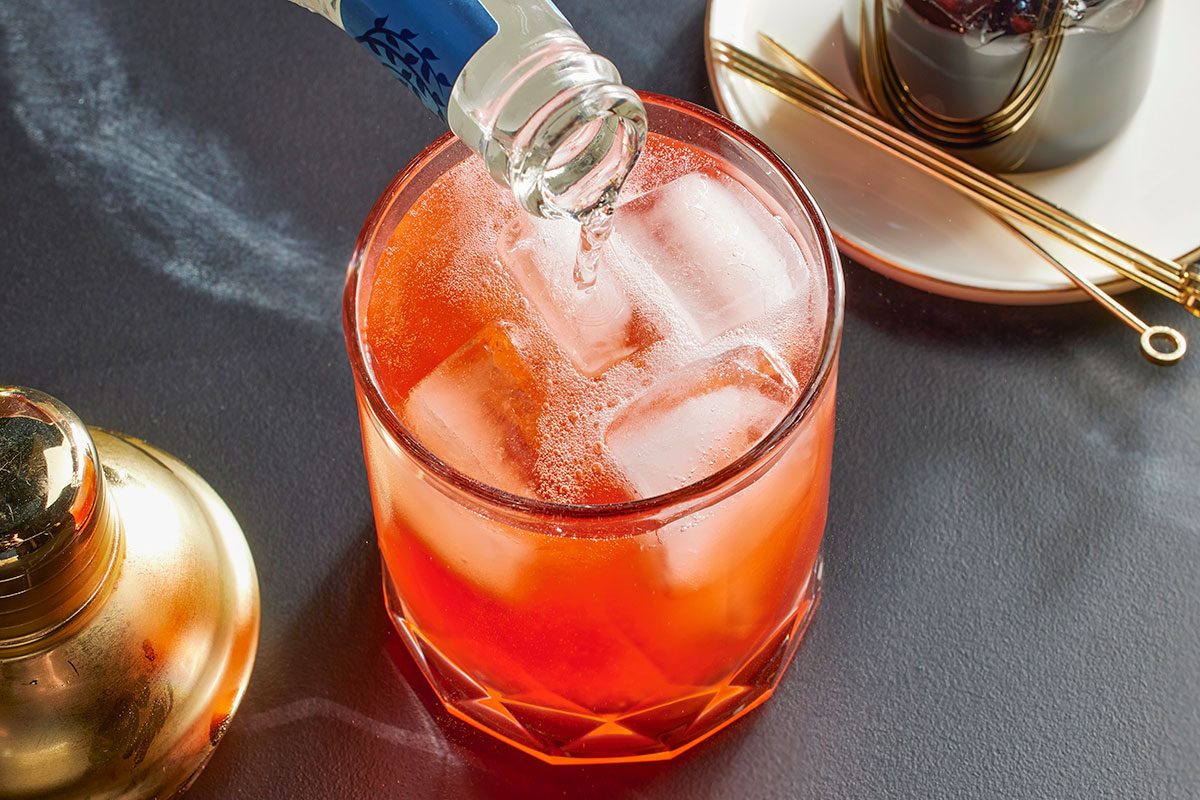 Overhead horizontal image of a glass filled with ice and a red liquid, likely a cocktail. A bottle is pouring clear liquid into the glass. A cocktail shaker and cocktail picks are visible to the side.