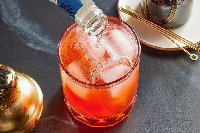 Overhead horizontal image of a glass filled with ice and a red liquid, likely a cocktail. A bottle is pouring clear liquid into the glass. A cocktail shaker and cocktail picks are visible to the side.