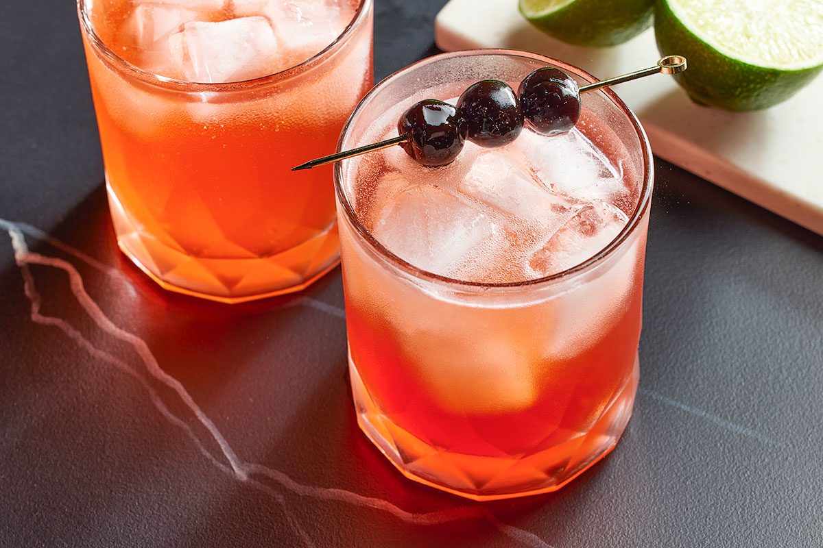 Eye Level horizontal image of two garnished cocktails in rocks glasses. The front drink has a cocktail pick of cherries and ice, limes are behind it.