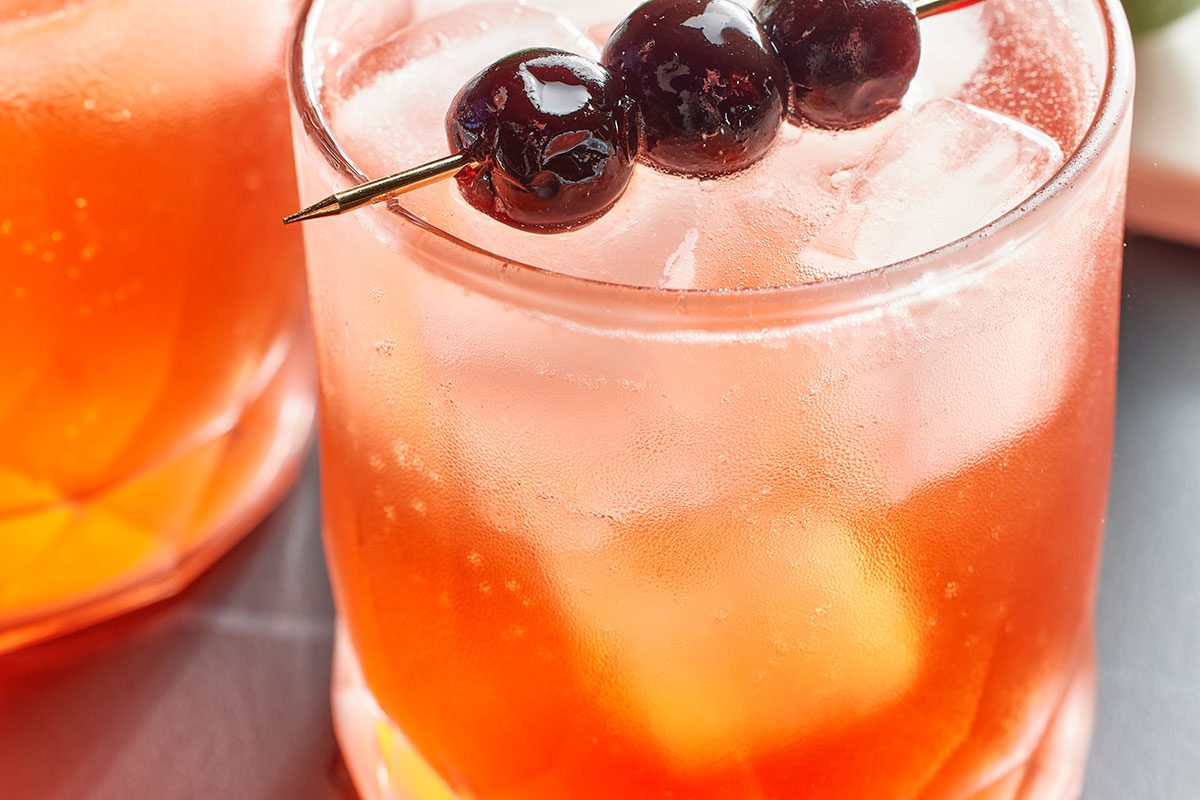 Eye Level horizontal image of a cocktail glass filled with an orange liquid and ice. The cocktail is garnished with three cherries on a toothpick, and the glass is slightly frosted.