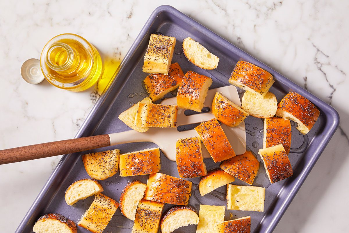 Overhead horizontal image of cut pieces of poppy seed bread and plain bread on a dark grey baking sheet, with visible drops of oil. A spatula is seen mixing the bread pieces, next to a bottle of cooking oil on a marble surface.