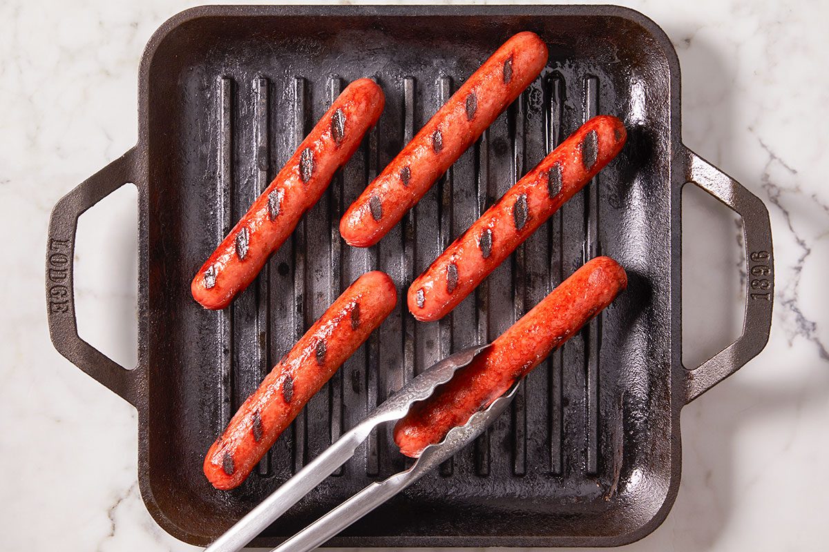 Overhead horizontal image of several hot dogs grilling in a black cast iron grill pan. One hot dog, showing distinct grill marks, is held by a pair of metal tongs, while others rest in the pan.