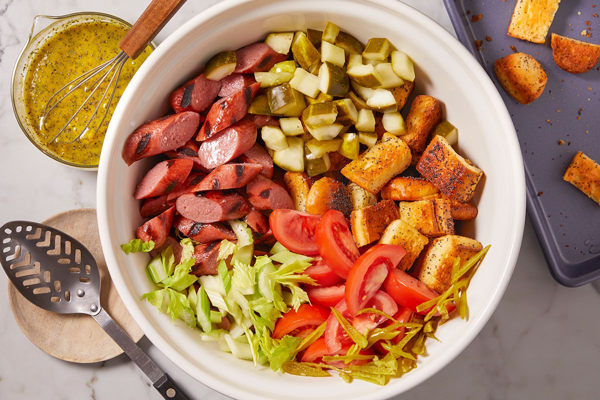 Overhead horizontal image of a large white bowl filled with ingredients for a colorful salad. The bowl contains sliced grilled sausage, diced pickles, seasoned croutons, sliced red tomatoes, chopped green celery, and thin strips of yellow-green pepperoncini. A glass measuring cup with a mustard-colored dressing and a whisk, along with a slotted spoon, are visible beside the bowl.
