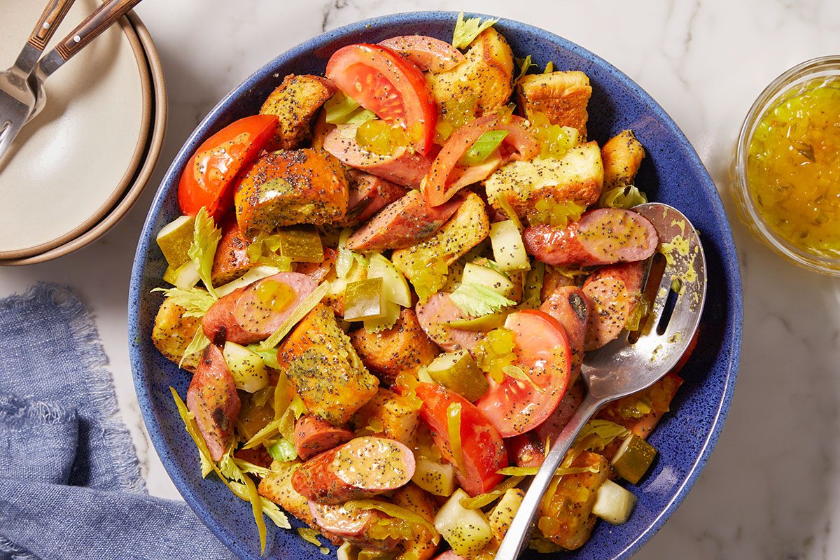 Overhead horizontal image of a hearty salad served in a blue bowl. The salad features sliced sausage, toasted bread cubes, quartered tomatoes, chopped celery, and diced pickles, all coated in a poppy seed dressing. A slotted serving spoon rests in the bowl, with an extra dish of dressing visible on the side.