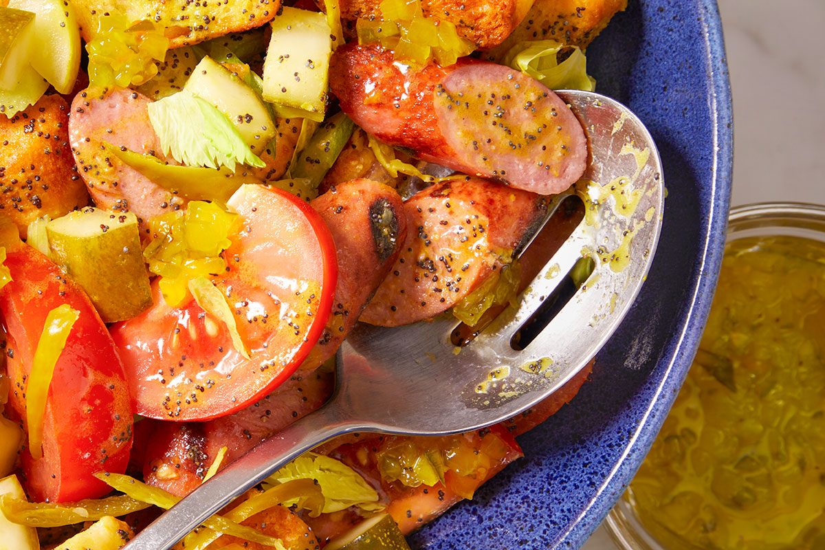 Overhead horizontal image of a hearty salad being mixed with a slotted spoon in a blue bowl. Sliced sausage, fresh tomato, pickles, celery, and croutons are tossed together and coated in a poppy seed dressing.