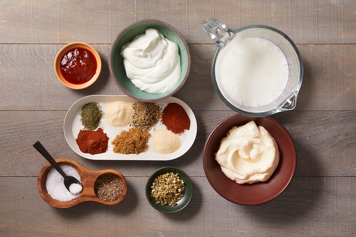 Assorted ingredients for a recipe, including bowls of sour cream, mayonnaise, milk, ketchup, and various spices and seasonings arranged on a wooden surface.