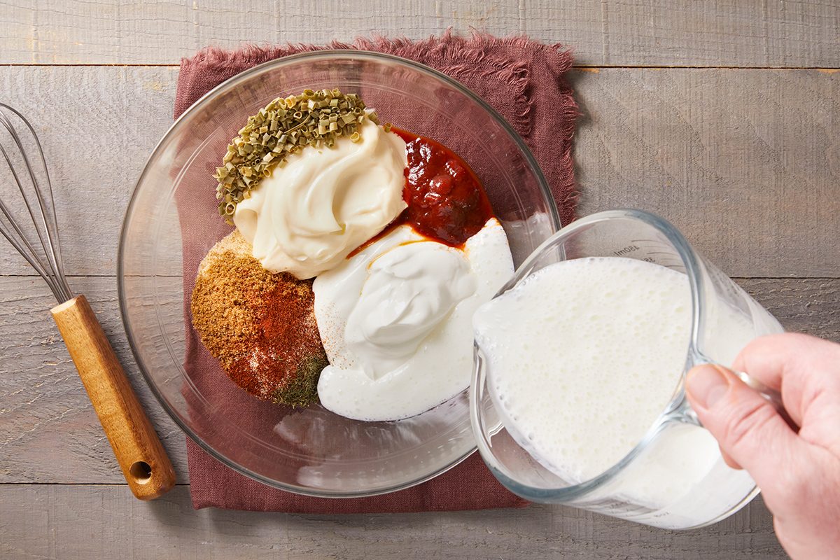 A hand pours a white liquid from a measuring cup into a glass bowl containing mayonnaise, ketchup, spices, and seasonings. A whisk rests on the table beside the bowl.