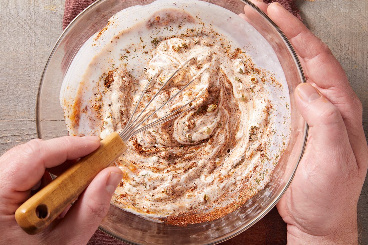 A person whisking a creamy mixture with brown spices in a clear glass bowl, viewed from above on a wooden surface.