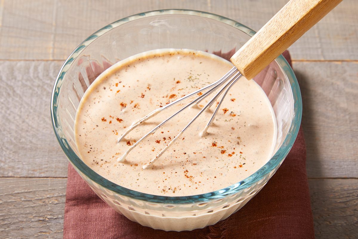 A glass bowl filled with creamy dressing, sprinkled with black pepper, sits on a folded cloth. A metal whisk with a wooden handle rests inside the bowl on a wooden surface.
