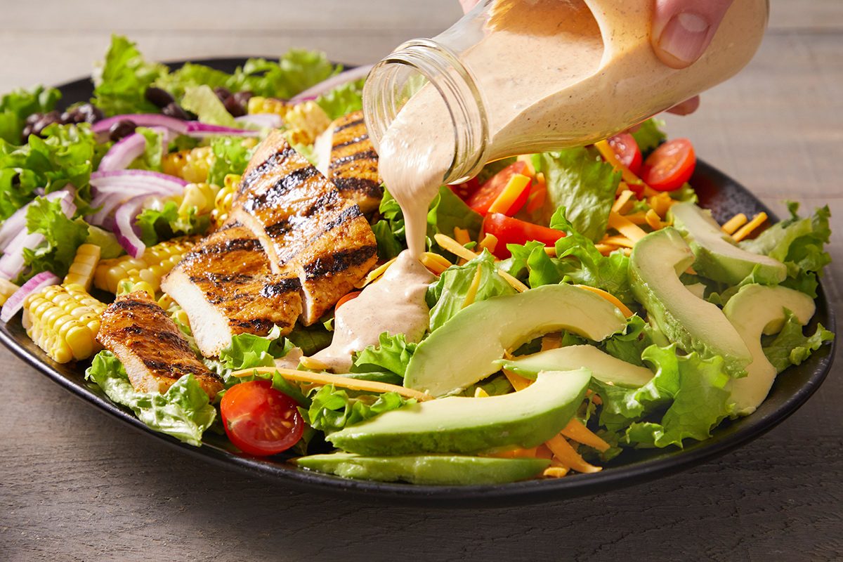 A hand pours creamy dressing over a salad with grilled chicken, avocado slices, cherry tomatoes, shredded cheese, red onions, corn, and mixed greens on a black plate.