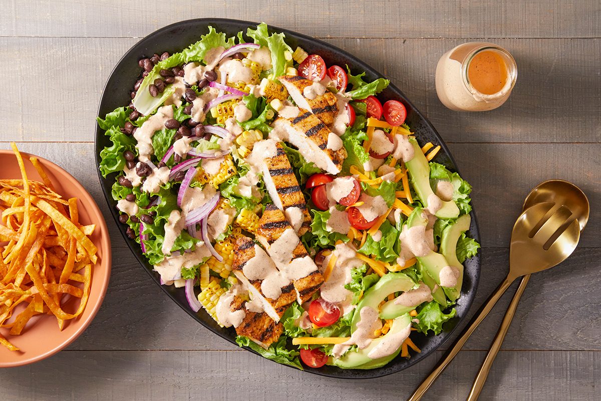 A large oval platter with grilled chicken salad featuring lettuce, cherry tomatoes, avocado slices, shredded cheese, red onions, and dressing, next to a bowl of crispy tortilla strips and two gold utensils.