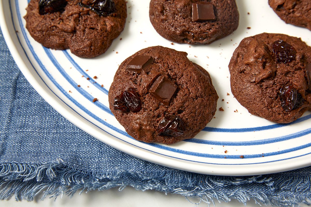 A plate with several chocolate cookies studded with chocolate chunks and dark dried fruit, placed on a blue denim-textured cloth.