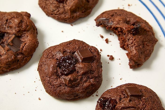 Five double chocolate cookies with visible chocolate chunks are on a white plate with blue lines. One cookie has a bite taken out of it, and there are small crumbs scattered around.