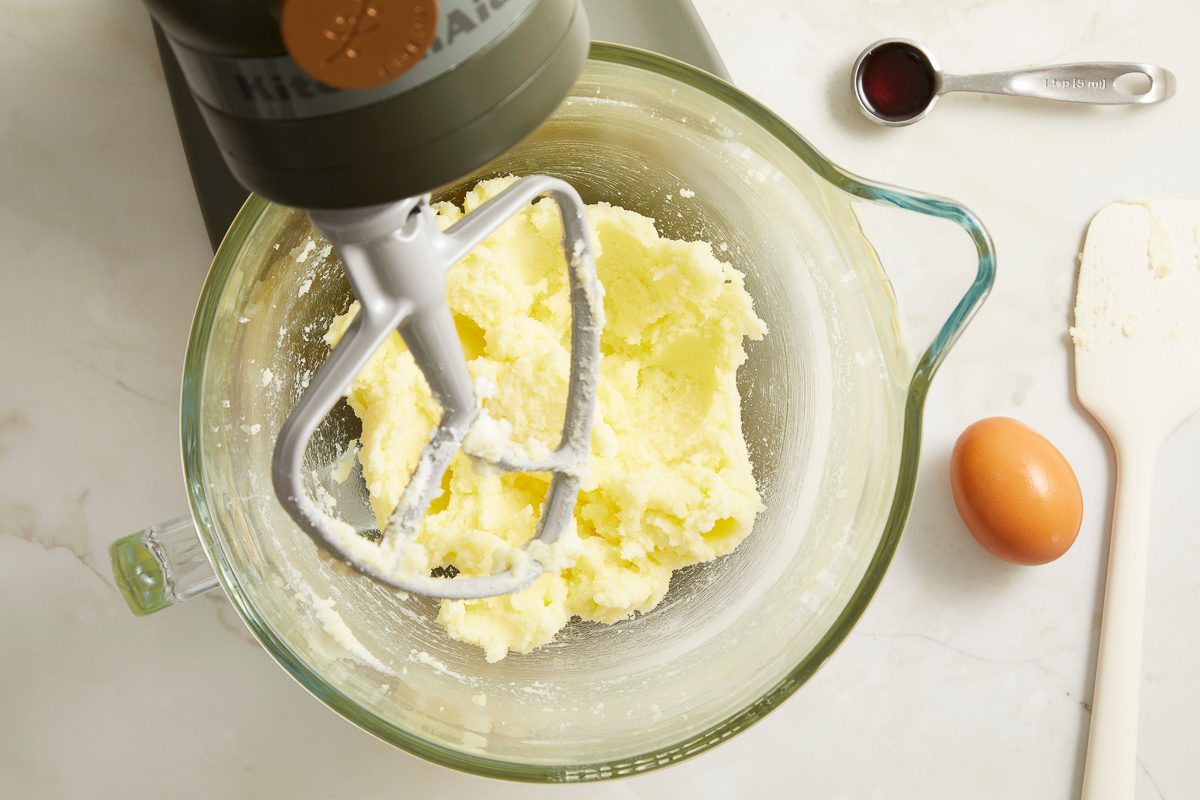 Overhead horizontal image of creamed butter and sugar in a glass mixing bowl, attached to a KitchenAid stand mixer. A measuring spoon filled with vanilla extract, a brown egg, and a rubber spatula are visible on the marble countertop next to the bowl.