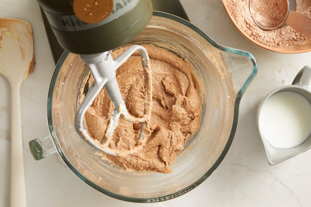 Overhead horizontal image of a KitchenAid stand mixer with a beater attachment still in a glass bowl, mixing a thick brown batter. To the right, a small pitcher of milk and a bowl of cocoa powder with a measuring cup are visible. A spatula with some batter on it rests to the left.