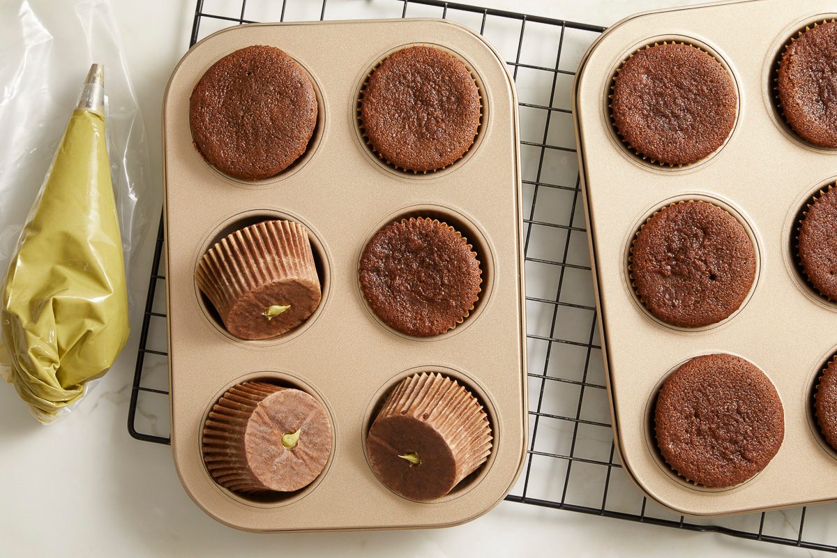 Overhead horizontal image of a gold muffin pan containing baked chocolate cupcakes. Some cupcakes have been cored and filled with a green frosting, with a piping bag filled with the same green frosting visible on the left.