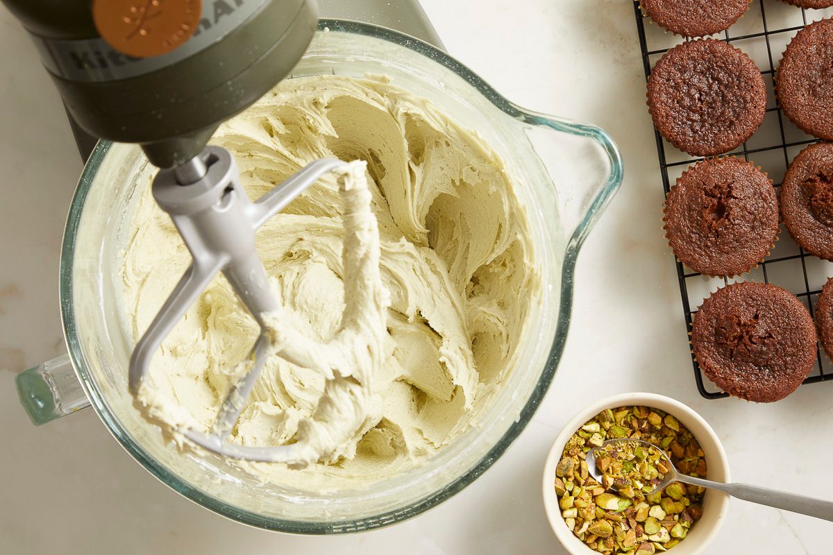 Overhead horizontal image of a stand mixer beating a light green frosting in a clear glass bowl. Next to the bowl, a cooling rack holds several chocolate cupcakes, and a small white bowl contains chopped pistachios with a spoon.