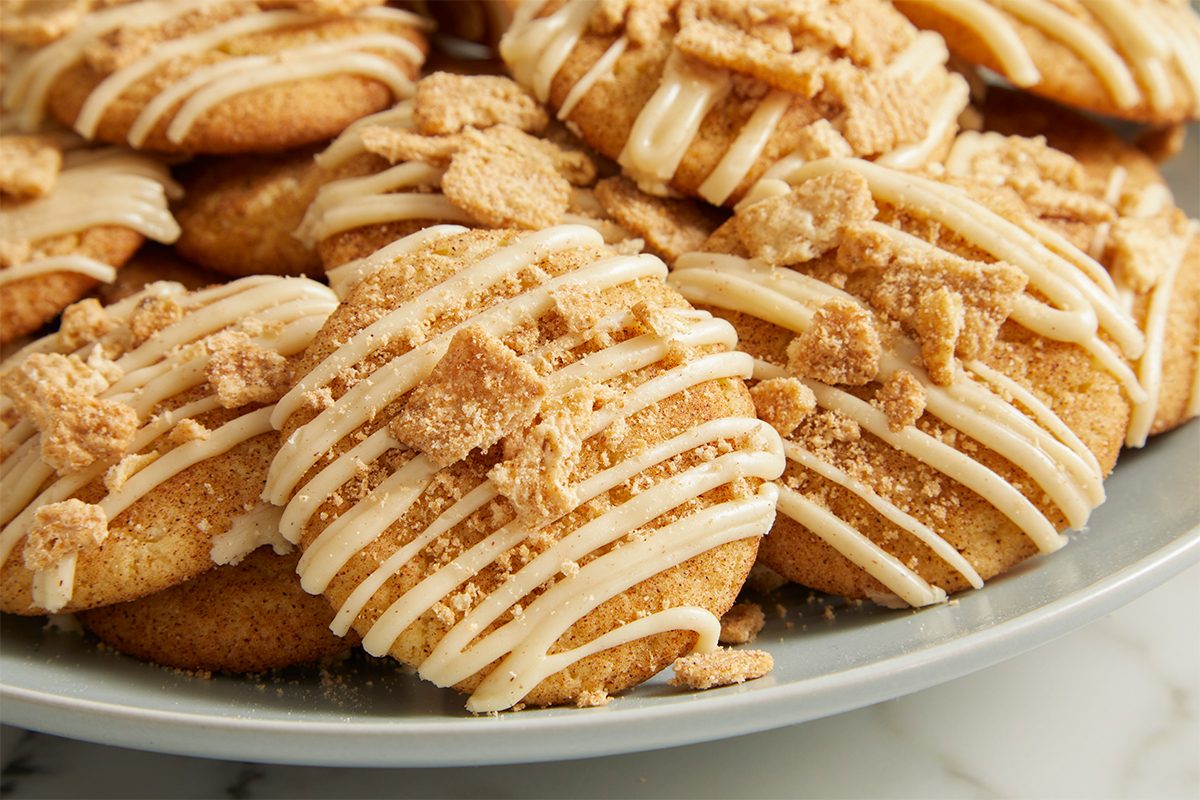 Eye Level horizontal image of a plate full of Snickerdoodle cookies. The cookies are topped with a white drizzle and chunks of cinnamon-flavored topping.