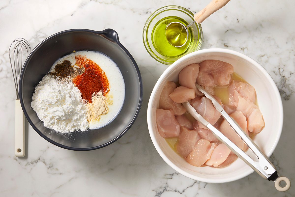 A horizontal overhead image shows raw chicken pieces in a bowl alongside measured ingredients including cornstarch, spices, and oil arranged on a countertop.