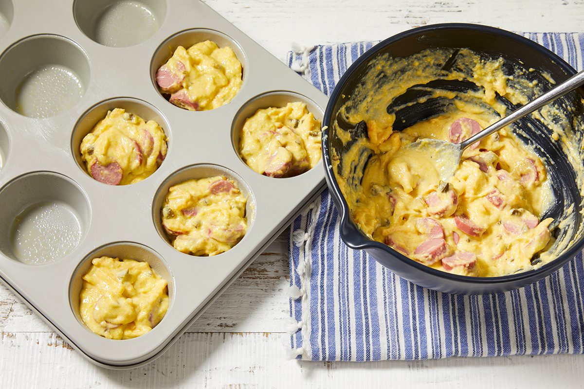 Overhead horizontal image of a muffin tin partially filled with Cheddar Corn Dog Muffin batter. A mixing bowl with the remaining batter, containing visible pieces of sliced turkey hot dogs, shredded sharp cheddar cheese, and chopped pickled jalapeno, sits next to it with a spoon.