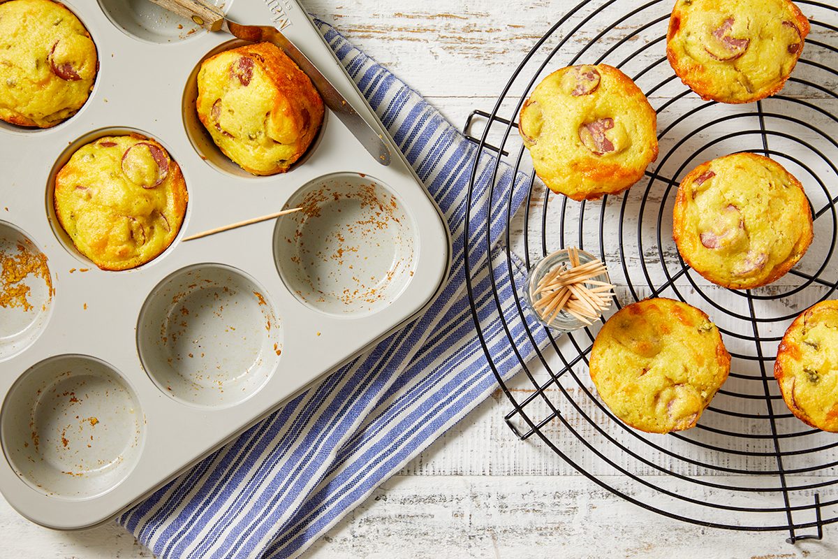 Overhead horizontal image of baked Cheddar Corn Dog Muffins being removed from a muffin tin. Several golden-brown muffins, dotted with turkey hot dog slices, are cooling on a wire rack next to the tin, while others remain in the pan, with one being tested by a toothpick.