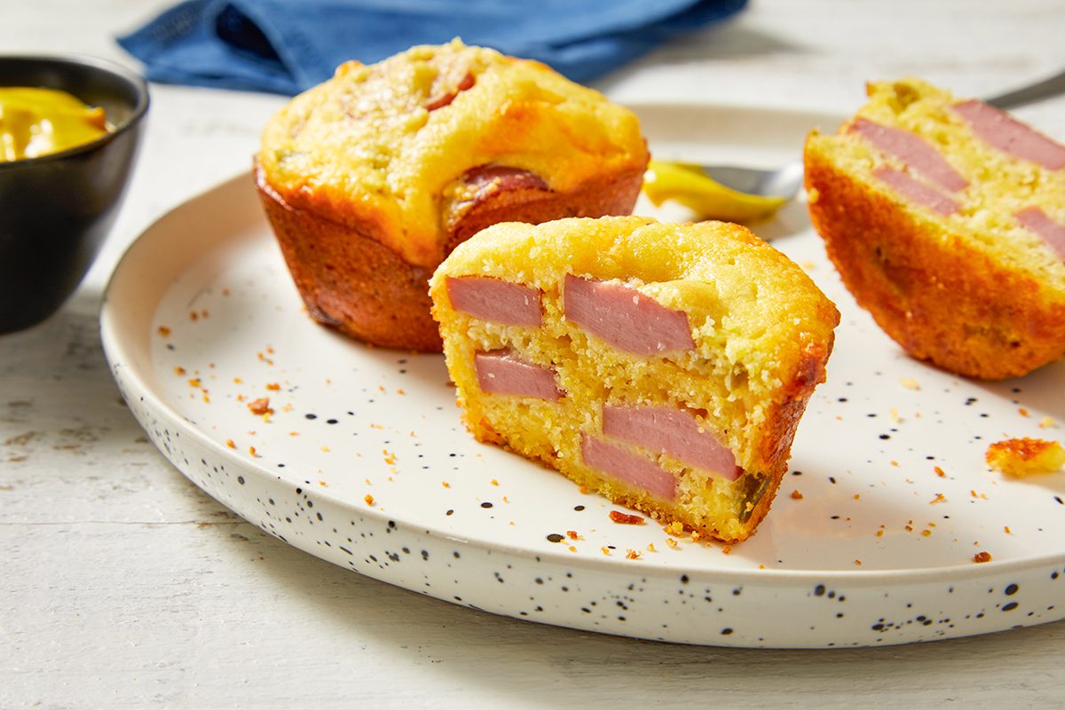 Eye Level horizontal image of three golden-brown Cheddar Corn Dog Muffins on a speckled white plate. One muffin is cut in half, revealing slices of turkey hot dogs and bits of cheddar cheese and pickled jalapeno. A small black bowl of mustard is visible in the background, ready for dipping.