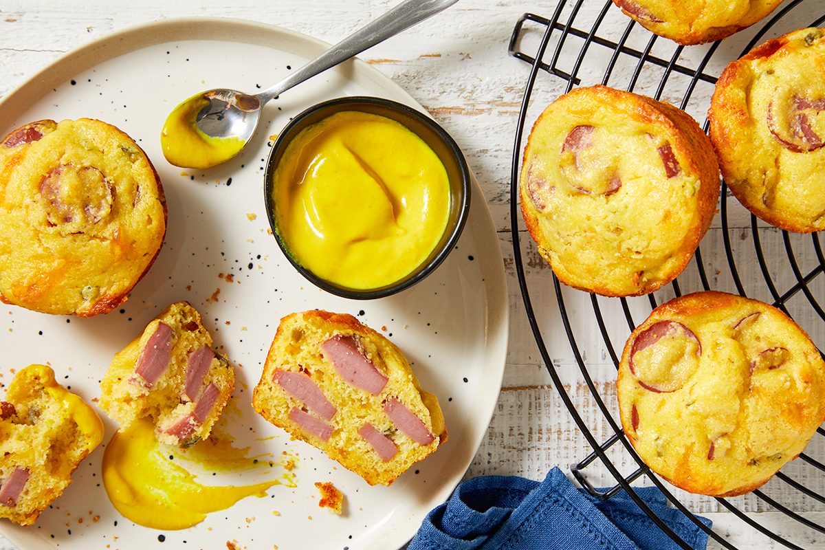 Overhead horizontal image of freshly baked Cheddar Corn Dog Muffins presented on a white plate and cooling rack. A small bowl of yellow mustard and a spoon are on the plate, with one muffin cut open to display the turkey hot dog pieces inside.
