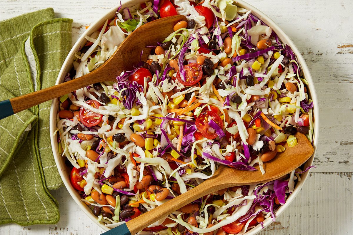 Overhead horizontal image of a vibrant, colorful salad in a large white bowl, featuring shredded green and red cabbage, halved cherry tomatoes, corn kernels, black beans, pinto beans, and shredded carrots. Two wooden serving spoons are resting in the salad, which is placed on a distressed white wooden surface next to a folded green kitchen towel.