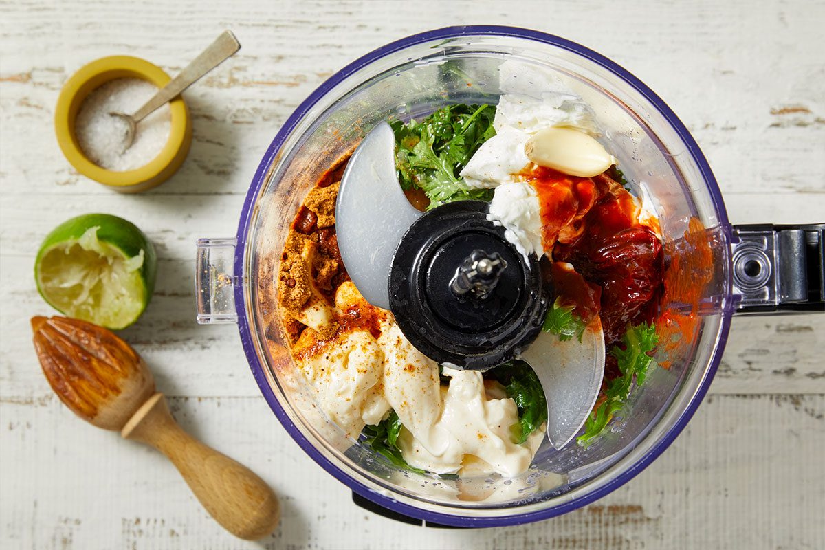 Overhead horizontal image of various ingredients loaded into a food processor bowl, ready to be blended. The bowl contains mayonnaise or sour cream, chipotle sauce, green herbs, powdered spices, and a whole garlic clove. A squeezed lime half, a wooden citrus reamer, and a small dish of salt are visible next to the processor on a white wooden surface.