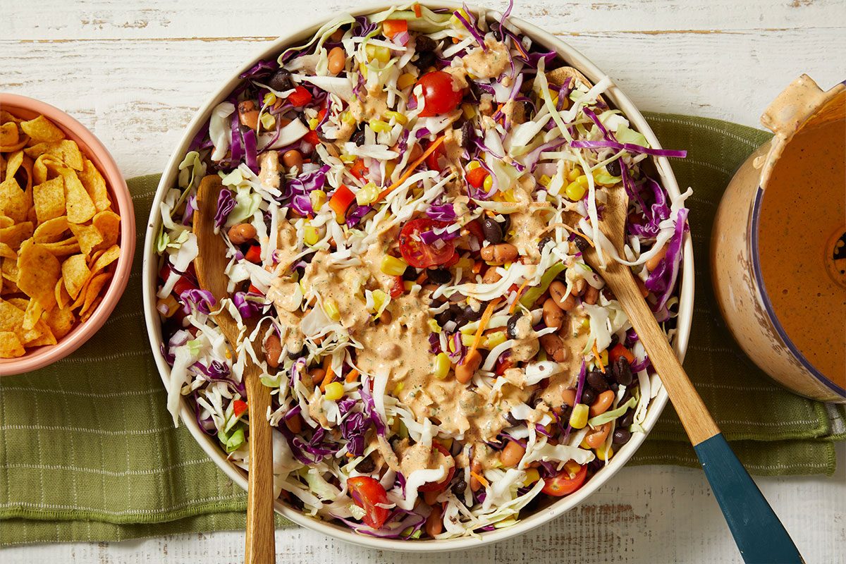 Overhead horizontal image of a large salad bowl filled with shredded green and red cabbage, corn, black beans, pinto beans, cherry tomatoes, red bell pepper, and shredded carrots, with a creamy dressing drizzled over it and a wooden serving spoon. A bowl of corn chips and a pitcher of dressing are visible on either side.
