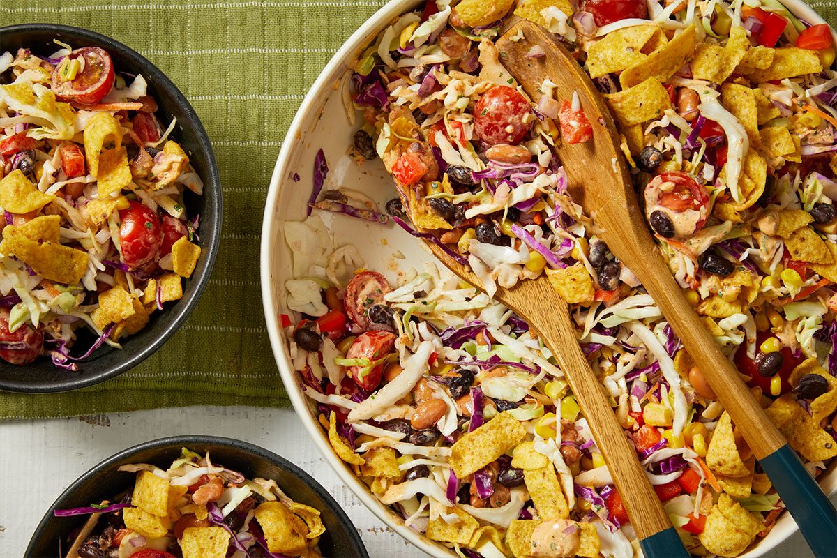 Overhead horizontal image of a vibrant Mexican-style salad. A large serving bowl brimming with shredded cabbage, black beans, pinto beans, corn, halved cherry tomatoes, and crispy corn chips, with two wooden salad servers, is positioned next to two individual black bowls also filled with the salad, all resting on a green textured mat and white surface.