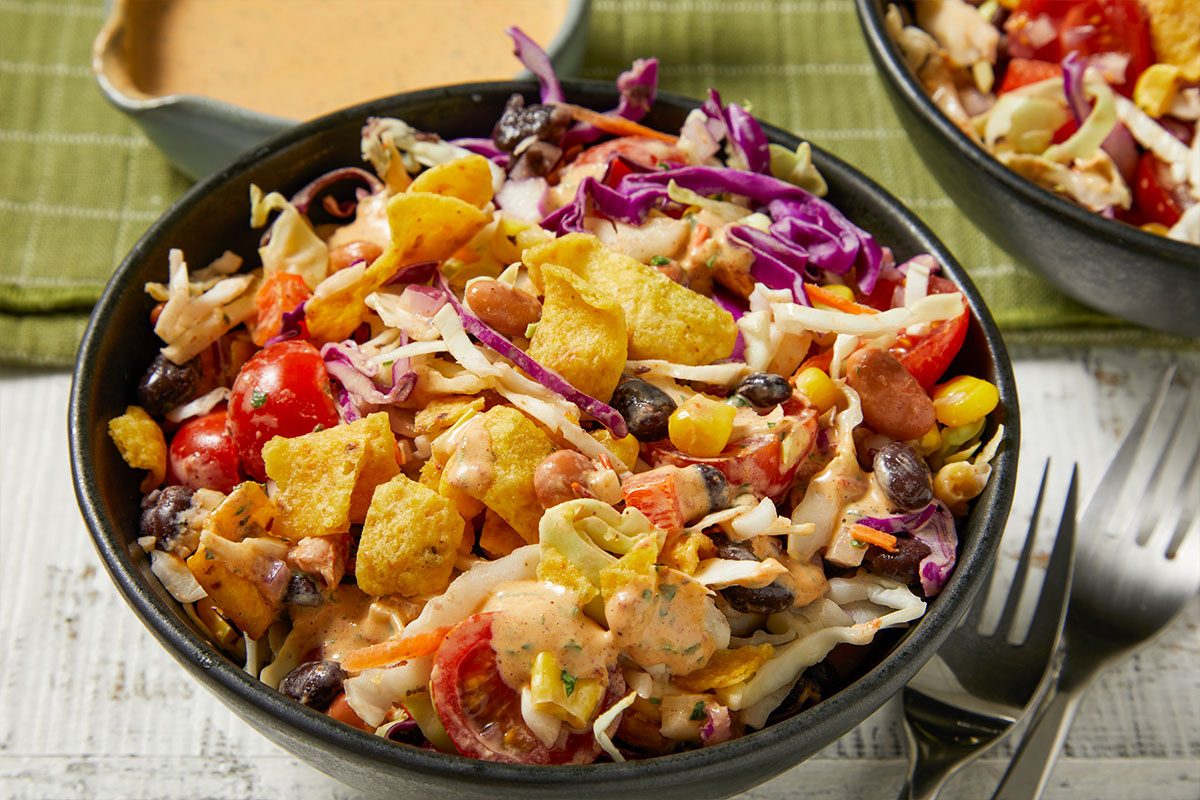 Eye Level horizontal image of a colorful salad served in a black bowl. The salad is composed of shredded white and red cabbage, black beans, corn, halved grape tomatoes, kidney beans, and crunchy corn chips, all mixed with a creamy dressing.