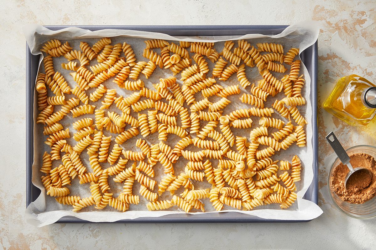Overhead horizontal image of fusilli pasta spread evenly on a parchment-lined baking sheet. The pasta is coated with a reddish-brown spice blend, and a bottle of cooking oil along with a bowl of spices and a measuring spoon are visible to the right.