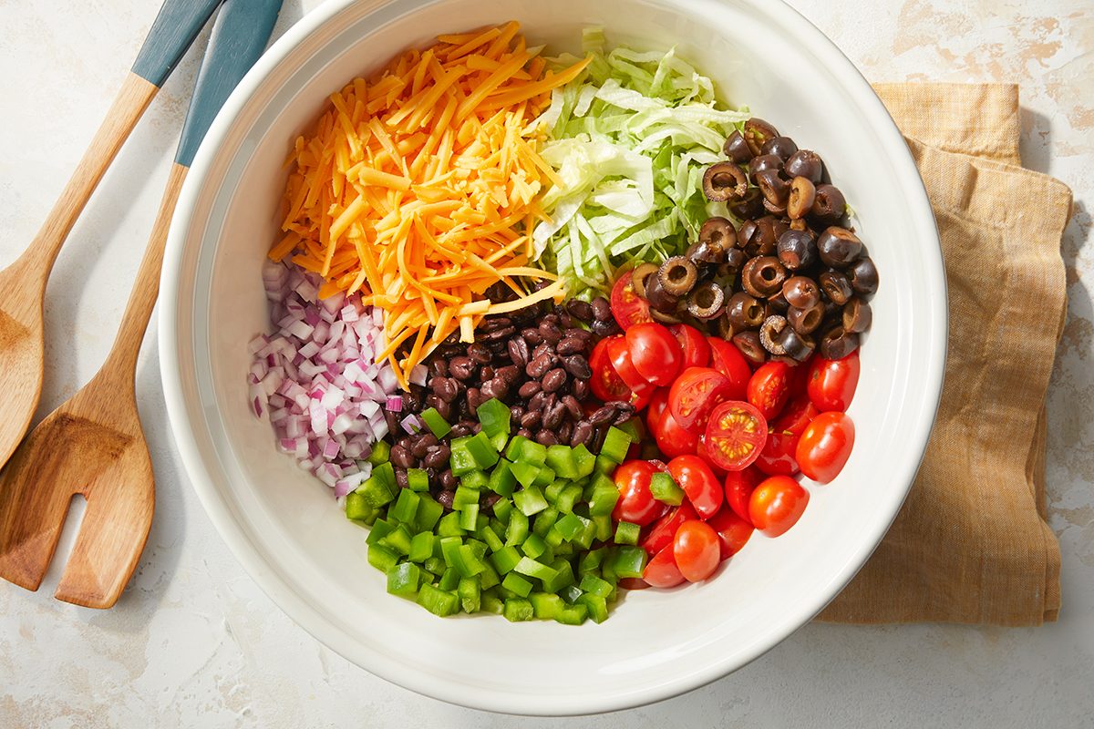 Overhead horizontal image of a large white bowl filled with assorted salad ingredients arranged in sections. The bowl contains shredded cheddar cheese, shredded lettuce, sliced black olives, halved cherry tomatoes, diced green bell pepper, black beans, and diced red onion. Wooden salad servers and a tan napkin are visible beside the bowl.