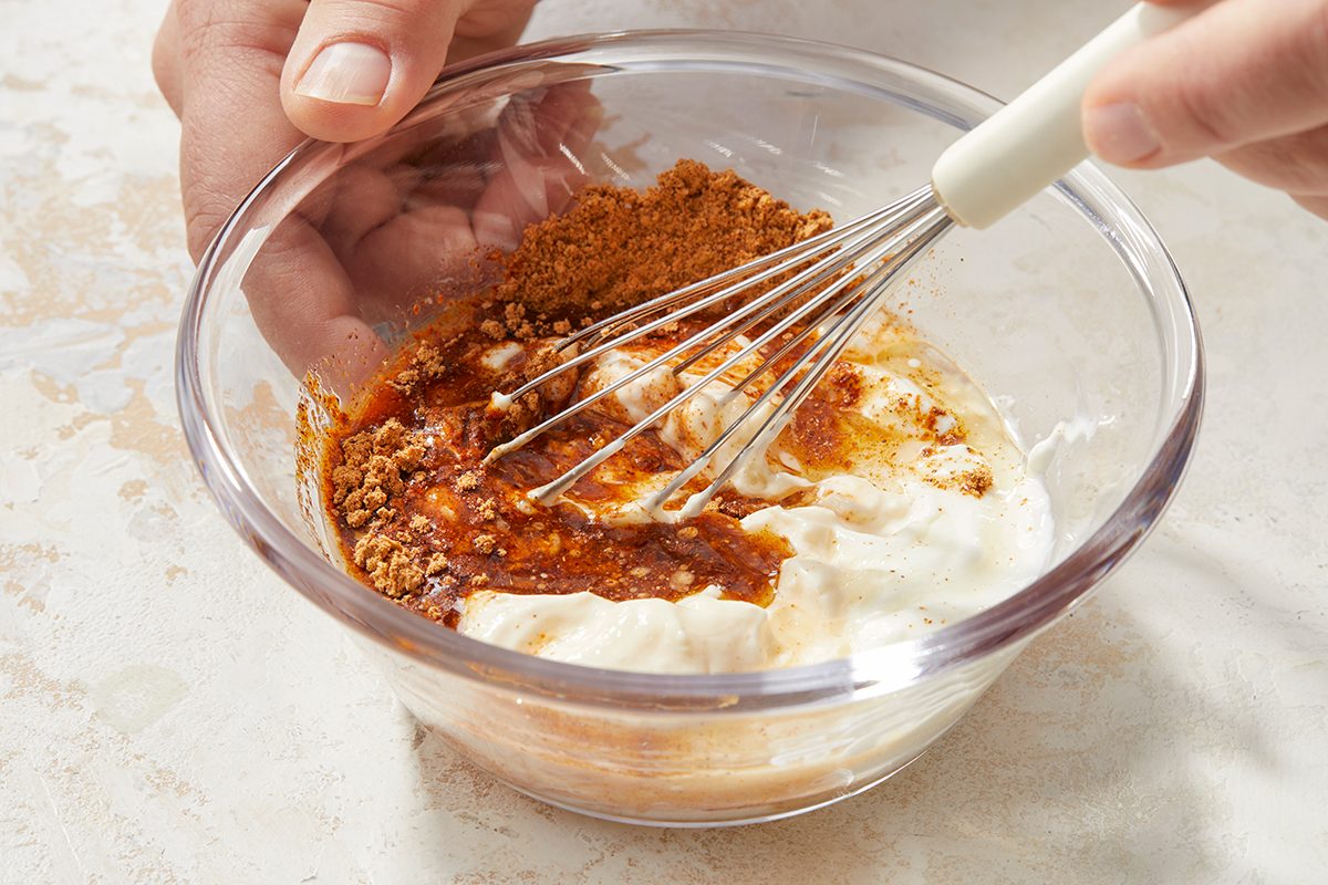 Eye Level horizontal image of hands holding a clear glass bowl. A whisk is positioned within the bowl, starting to mix a creamy white base with brown spices and a dark liquid, indicating the beginning of a blending process.