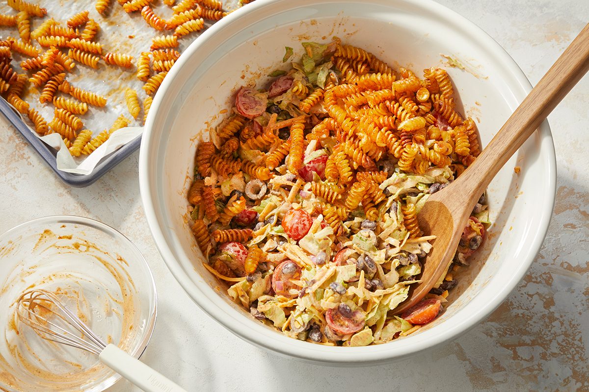 Overhead horizontal image of golden-brown rotini pasta being added to or mixed into a creamy salad in a large white mixing bowl. The salad contains shredded cabbage or lettuce, halved cherry tomatoes, black beans, and sliced black olives, coated in a creamy dressing. A baking sheet with more roasted rotini pasta is visible in the background, and an empty whisk and bowl are in the foreground.