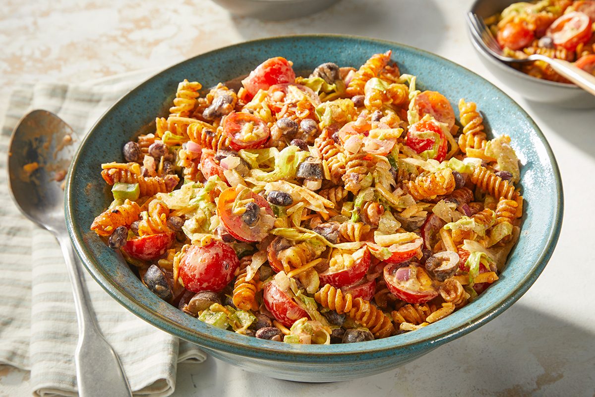 Eye level horizontal image of a large blue bowl filled with a vibrant pasta salad. The salad features spiral pasta, halved cherry tomatoes, black beans, sliced black olives, shredded lettuce, and red onion, coated in a creamy dressing. A spoon rests beside the bowl on a striped napkin, with another small bowl of the salad in the soft-focus background.