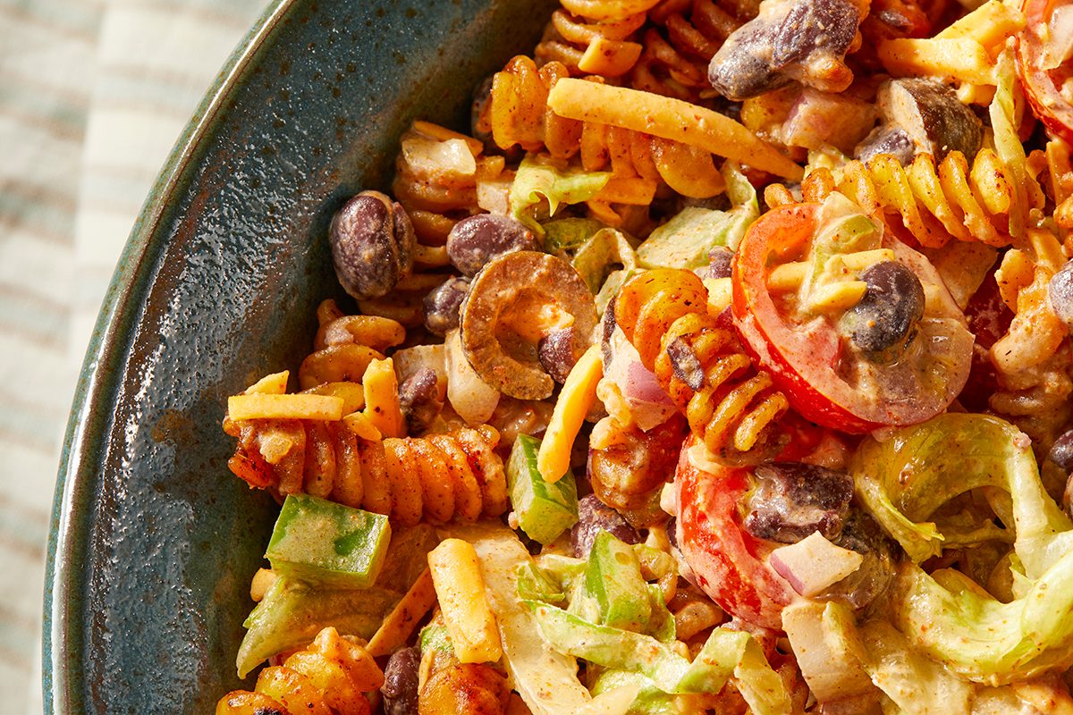 Overhead horizontal image of a vibrant pasta salad served in a blue ceramic bowl. The salad features spiral pasta coated in a creamy dressing, mixed with black beans, sliced tomatoes, red onion, green bell peppers, and sliced black olives. Hints of shredded lettuce are also visible, adding a fresh texture to the dish.