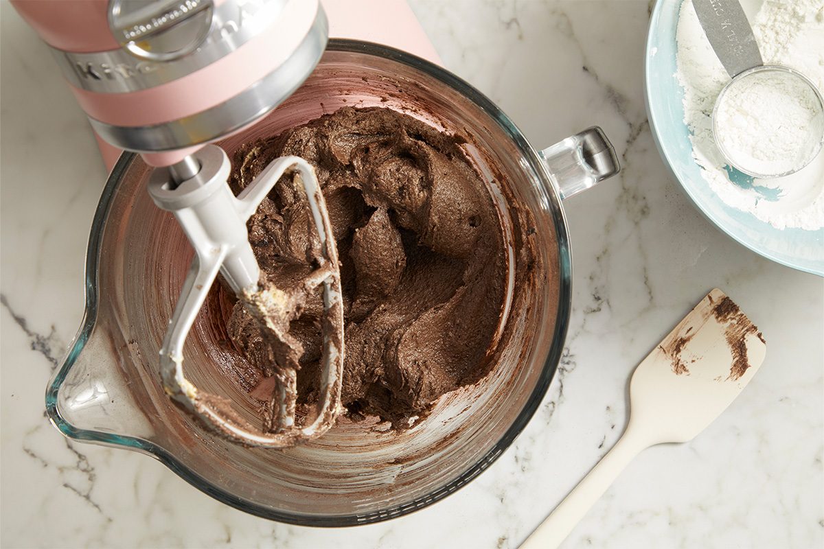 Overhead horizontal image of a pink KitchenAid stand mixer actively mixing dark brown batter in a clear glass bowl with a flat beater attachment. A light blue bowl containing white flour and a measuring cup, along with a white spatula with some batter on it, are visible on the marble countertop next to the mixer.