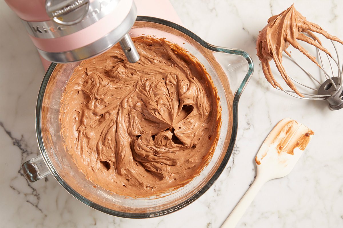 Overhead horizontal image of a pink stand mixer whisking creamy chocolate frosting in a clear glass measuring cup. A metal whisk coated in the rich chocolate frosting rests on the white marble countertop, alongside a spatula with some frosting residue.