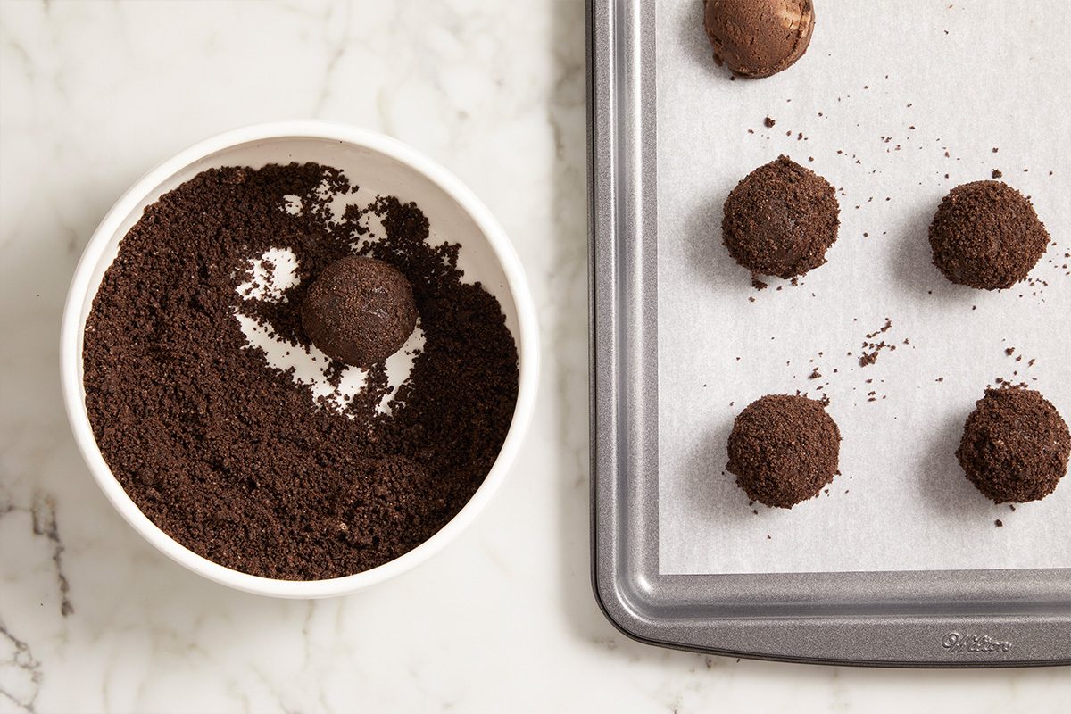 Overhead horizontal image of a dark brown ball being coated in a white bowl filled with dark brown cookie crumbs. To the right, a baking sheet lined with parchment paper holds several other crumb-coated dark chocolate balls and one uncoated ball, ready for further preparation.