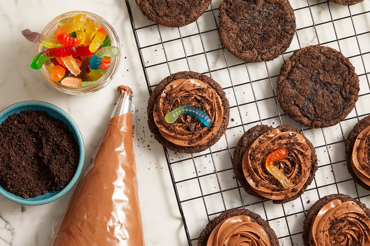 Overhead horizontal image of chocolate cookies being decorated. Several chocolate cookies with chocolate frosting are topped with cookie crumbs and colorful gummy worms, while a piping bag with frosting, a bowl of cookie crumbs, and a jar of gummy worms sit nearby on a marble surface.