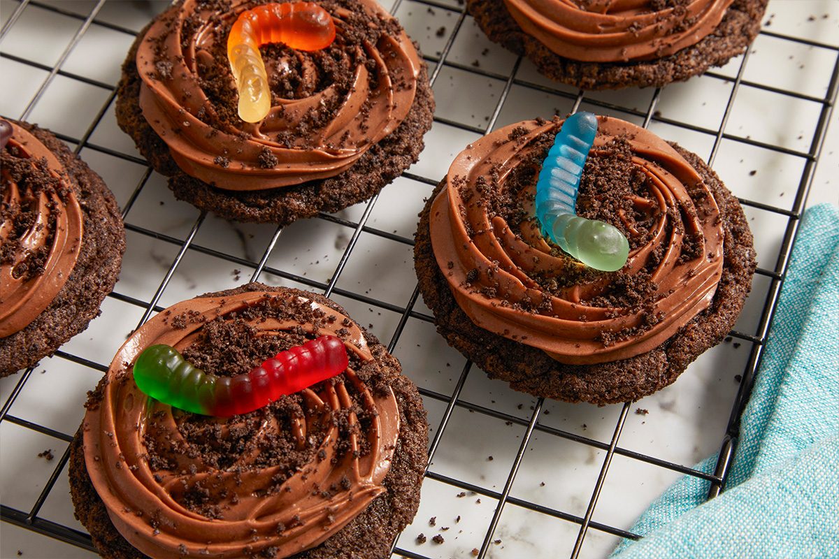 Eye Level horizontal image of four chocolate cookies with rich chocolate frosting, sprinkled with crushed cookie crumbs, and adorned with colorful gummy worms. The cookies rest on a black wire cooling rack, with a light blue cloth visible to the right and a white marble surface underneath.