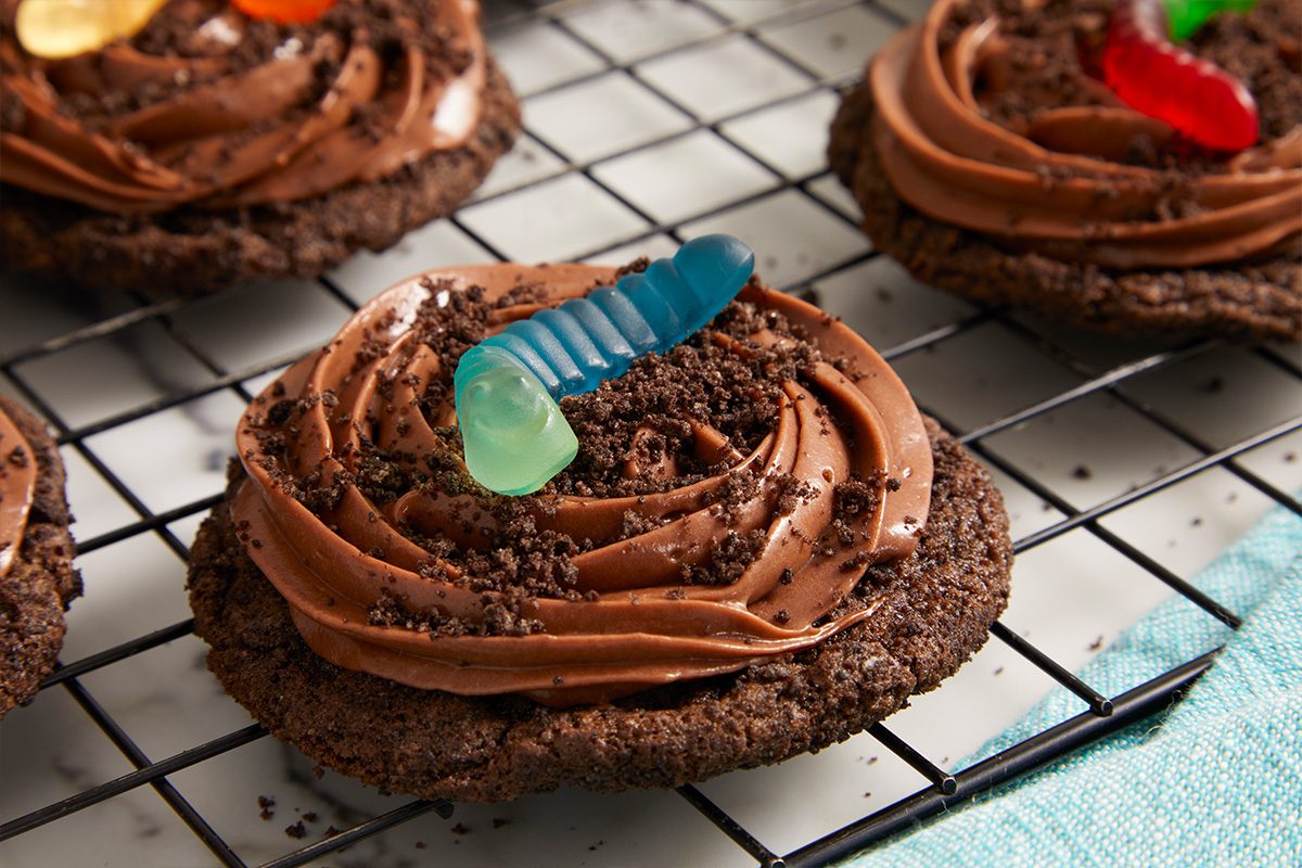 Eye Level horizontal image of chocolate dirt pudding cookies topped with chocolate frosting, cookie crumbs, and colorful gummy worms, resting on a black wire cooling rack with a light blue linen nearby.