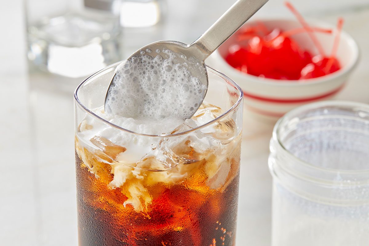 Eye Level horizontal image of a metal spoon scooping frothy white foam into a tall glass of iced dark beverage. The drink contains ice and swirls of cream, with maraschino cherries visible in a small bowl in the background.