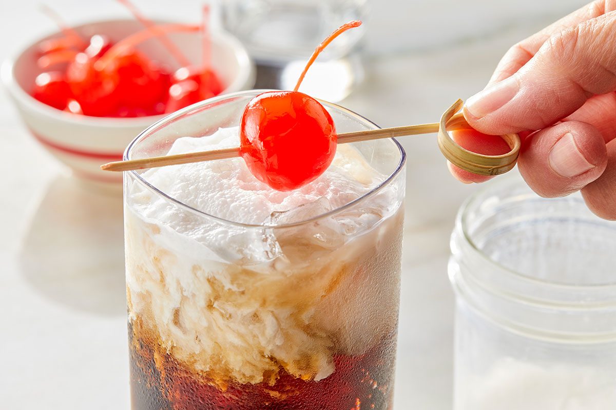 Eye Level horizontal image of a hand placing a maraschino cherry, skewered on a bamboo pick, onto a layered beverage in a tall glass. The drink features a dark liquid base, a lighter creamy middle layer, and a white whipped topping, with ice visible. A bowl of additional maraschino cherries sits blurred in the background.