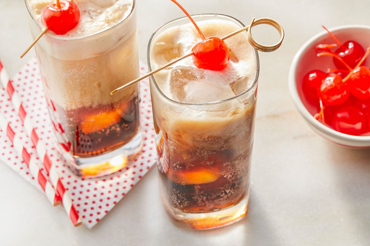 Eye Level horizontal image of two tall glasses of dark soda topped with creamy ice cream and ice cubes, each garnished with a bright red maraschino cherry on a skewer. A small white bowl of additional maraschino cherries and red and white striped paper straws are also visible in the frame.