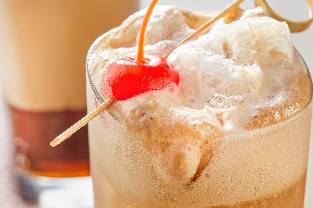 Eye Level horizontal image of a refreshing beverage in a clear glass, topped with foamy ice cream. A bright red maraschino cherry on a stick is nestled in the foam, which is slightly overflowing the glass.