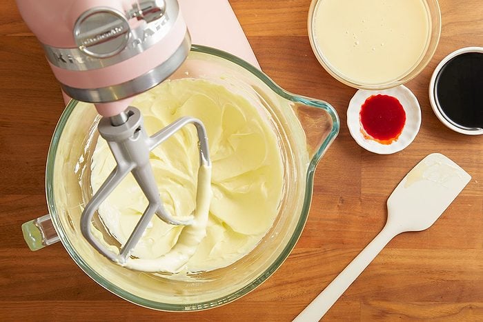 Overhead horizontal image of a pink KitchenAid stand mixer with a whisk attachment mixing a creamy yellow batter in a glass bowl. Alongside the bowl, there are separate small dishes containing light yellow liquid, dark liquid, and red food coloring, with a white spatula resting on the wooden surface.